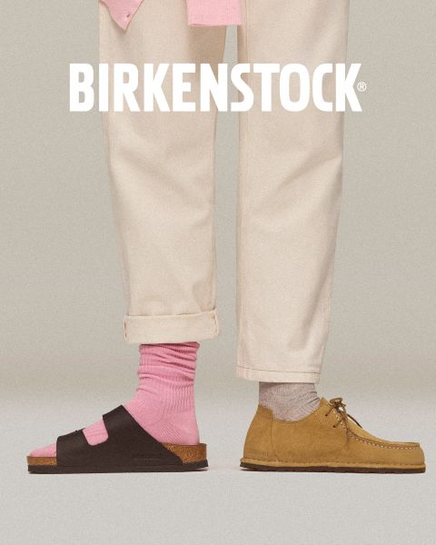 Un par de zuecos Birkenstock color beige con correas de hebilla sobre un suelo de ladrillo rojo junto a una pared de azulejos rojos con diseño geométrico. El logotipo blanco de Birkenstock está en la parte inferior de la imagen.