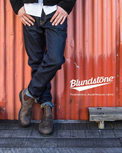 Una persona con botas Blundstone marrones desgastadas está de pie en un barco desgastado cerca de cuerdas, con agua y rocas borrosas al fondo.