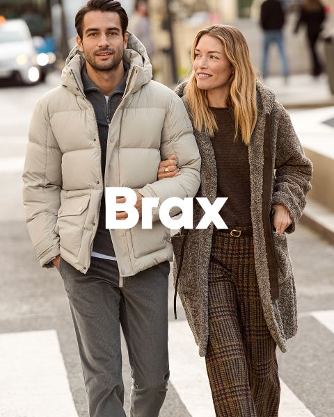Una mujer con un abrigo estampado y un suéter rojo está de pie junto a un hombre con un abrigo de invierno azul marino y pantalones beige. Están apoyados en una pared de piedra al aire libre. La palabra "Brax" aparece en la parte inferior de la imagen.