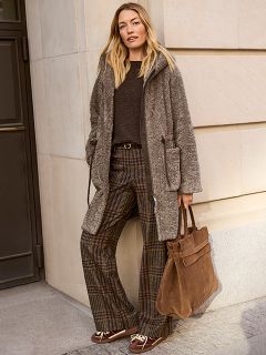 Una mujer con el pelo largo está de pie en una acera de la ciudad. Lleva un suéter negro con rayas blancas y pantalones negros de pierna ancha, y sostiene un bolso de mano negro grande mientras la luz del sol la ilumina desde atrás.