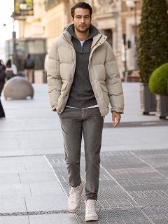 Un hombre con cabello corto y oscuro y barba está apoyado en una barandilla de piedra al aire libre. Lleva una chaqueta a cuadros roja y blanca sobre un suéter color crema y jeans azules. En el fondo se ven edificios urbanos.