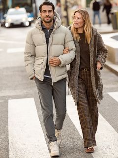 Una mujer con un abrigo de piel estampado y una blusa roja está de pie sonriendo junto a un hombre con una chaqueta de invierno azul marino y un suéter beige. Están de pie afuera junto a una pared de piedra, y la mujer sostiene un bolso marrón.