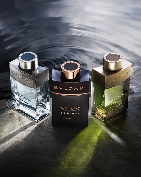 Una mujer con un vestido blanco se reclina frente a un fondo arquitectónico moderno bajo un cielo azul. En primer plano, dos frascos del perfume Bvlgari Omnia Crystalline, que muestran tonos claros y ámbar.