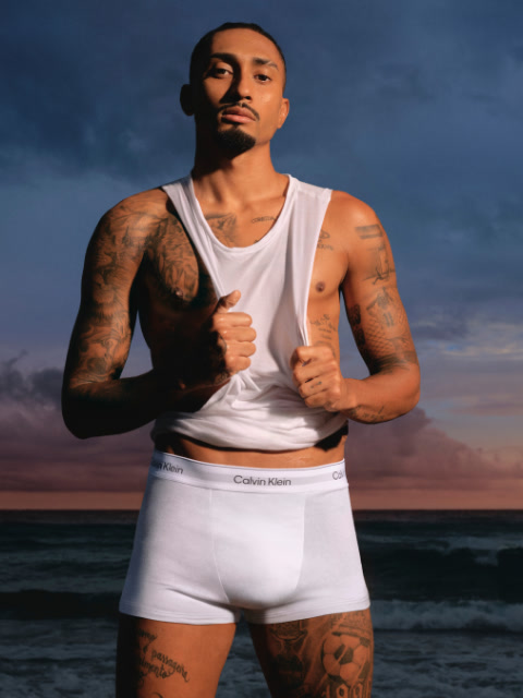 Un hombre con una chaqueta negra y vaqueros está de pie en un estadio y mira hacia un lado con las manos en los bolsillos. En el fondo se ven asientos de estadio y un gran edificio.