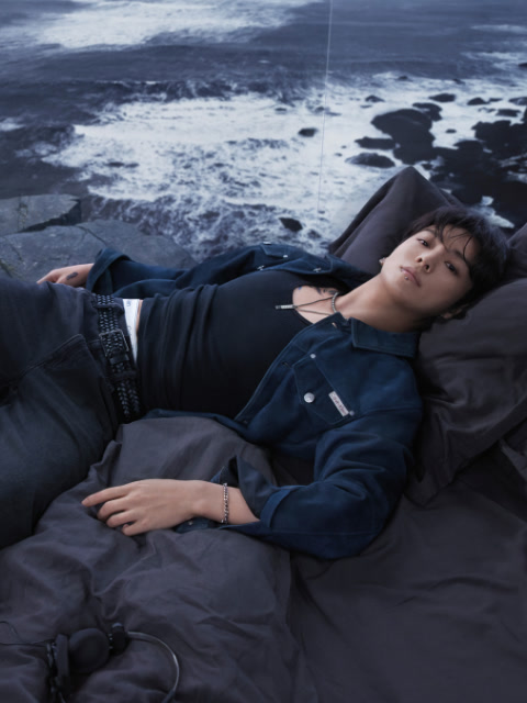 Fotografía en blanco y negro de una mujer con un sujetador deportivo de Calvin Klein y jeans, acostada boca arriba con las piernas dobladas sobre la cabeza, los brazos extendidos en el suelo y mirando a la cámara.