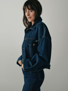 Una persona con cabello largo y rubio está sentada en un banco de madera, vistiendo una chaqueta vaquera azul, una camisa blanca y vaqueros de corte ancho. En el fondo se ven edificios urbanos y rascacielos.