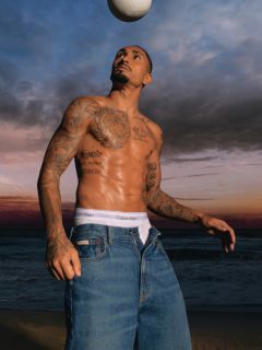 Un hombre con una chaqueta vaquera estampada y vaqueros azules está de pie en un puente con barandilla metálica. Al fondo se ven un río y otro puente en un día nublado.