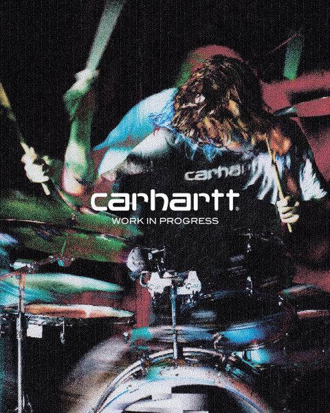 Imagen en blanco y negro de una persona con sombrero y chaqueta, agachada con las manos sobre las rodillas. El texto "Carhartt Work In Progress" se ve prominentemente en el centro.