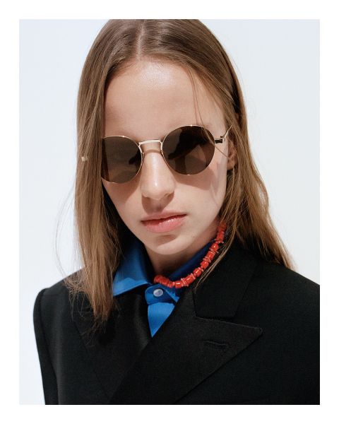 Fotografía en blanco y negro de una persona con cabello corto, gafas de sol oscuras y una camisa con cuello debajo de una chaqueta texturizada, mirando hacia un lado en un entorno con poca luz.