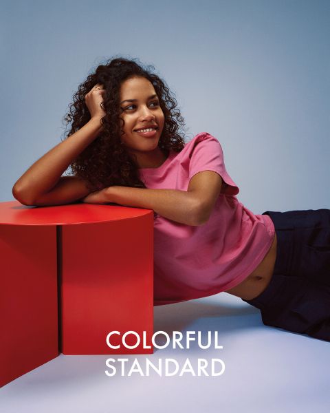 Una persona con cabello largo y rubio está de pie de lado frente a un fondo azul. Lleva una camiseta azul oscuro, vaqueros azul claro y zapatillas de deporte. El texto "COLORFUL STANDARD" se muestra en blanco.