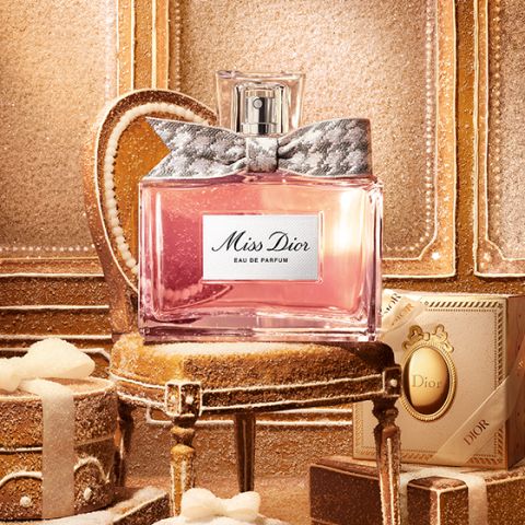 Una botella de cristal transparente de "Miss Dior Eau de Parfum", llena de líquido rosa y con un lazo plateado estampado alrededor del cuello de la botella, sobre un fondo blanco liso.