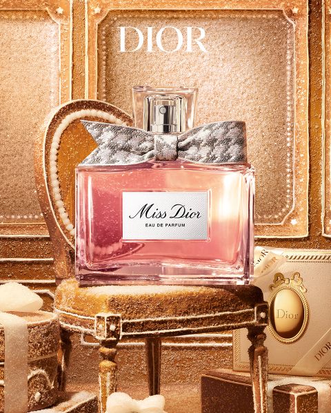 Un hombre con tatuajes y joyas se apoya en una mesa junto a una botella de perfume "Dior Sauvage Extrait" frente a un telón de fondo exterior rugoso.