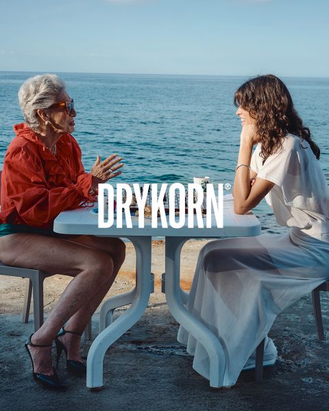 Una elegante mujer mayor con cabello rubio platino y un abrigo de piel negro está sentada en un sofá antiguo junto a un perro grande marrón. La palabra "DRYKORN" se puede leer en la parte inferior de la imagen. La iluminación cálida crea una atmósfera lujosa.