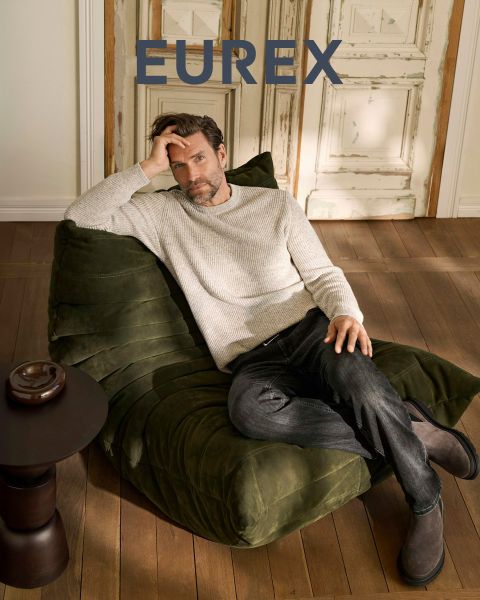 Un hombre está de pie al aire libre cerca de hierba alta, lleva un jersey beige, un chaleco acolchado azul marino y pantalones marrones, y mira hacia un lado. El logotipo de "EUREX by BRAX" aparece en el lado derecho. El cielo está nublado.