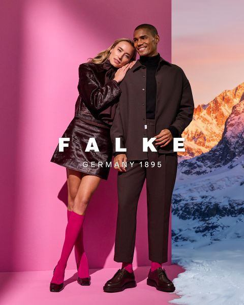 Un modelo con un blazer color castaño, guantes marrones, medias verdes, una falda gris con aberturas altas y botines marrones posa frente a un fondo geométrico. El texto dice "FALKE ALEMANIA 1895".