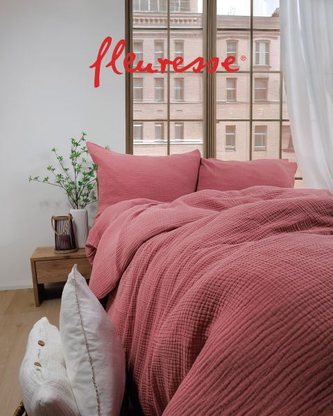 Un dormitorio acogedor con una cama bien hecha con ropa de cama blanca con un diseño de pájaros. Dos almohadas decorativas están apoyadas en el cabecero. La luz del sol entra a través de cortinas transparentes, proyectando un brillo suave sobre el suelo de madera clara.