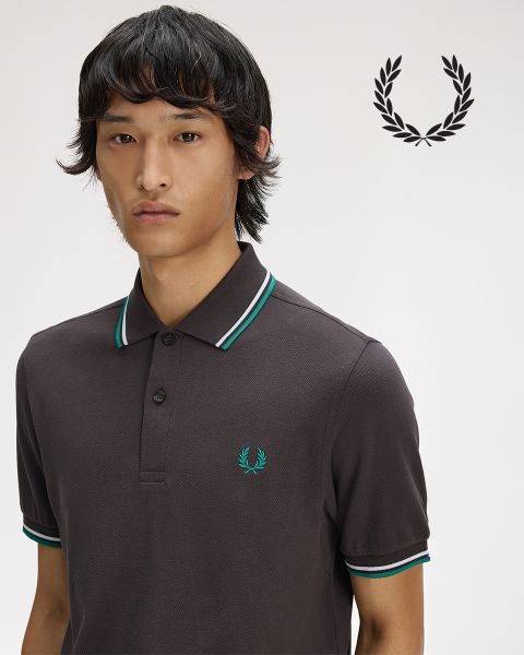 Una persona con cabello largo trenzado mira pensativamente hacia arriba mientras se ajusta una camisa blanca con cuello. La camisa está adornada con una corona de laurel. La imagen es en blanco y negro.