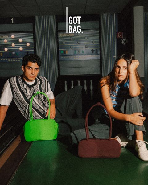 Hombre con gafas de sol sosteniendo grandes bolsas grises frente a un fondo a rayas oscuras. Debajo de él hay varias bolsas de diferentes tamaños y colores, todas expuestas ordenadamente. El logo en la esquina superior derecha dice "GOT BAG".