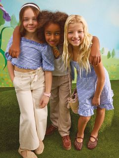 Tres jóvenes están de pie con confianza entre hojas de otoño, vistiendo atuendos brillantes y metálicos. Dos chicas llevan chaquetas plateadas, y la chica del centro lleva un top y una falda con lentejuelas. Un fondo verde azulado contrasta con el follaje naranja del otoño.