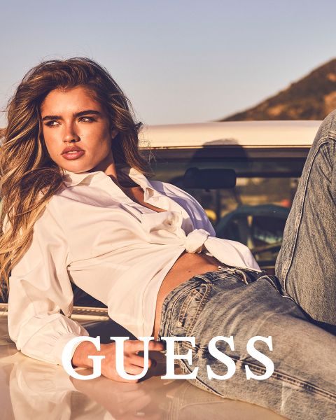 Dos mujeres están de pie juntas al aire libre, vistiendo abrigos texturizados, una en color canela claro y otra en color crema. Ambas sostienen sus cuellos y miran hacia adelante. La palabra "GUESS" está en negrita en letras blancas en la parte inferior.