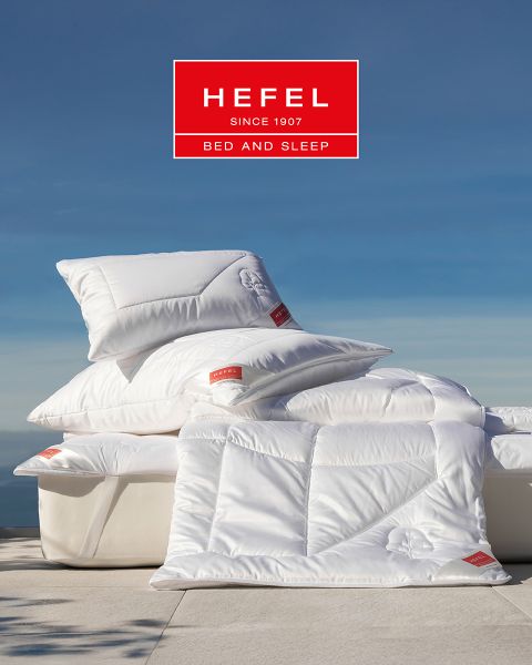 Una mujer duerme plácidamente de lado en una cama moderna con una almohada beige y una manta blanca. En el fondo, se ven un jarrón con hierba de la pampa y una vista de la montaña al atardecer. En la parte superior, se muestra el logotipo de "Hefel Bed and Sleep".