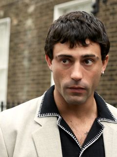 Una persona con un traje marrón y una gorra a juego está sentada en un banco negro frente a un fondo rojo y blanco, mirando hacia un lado con una expresión pensativa.