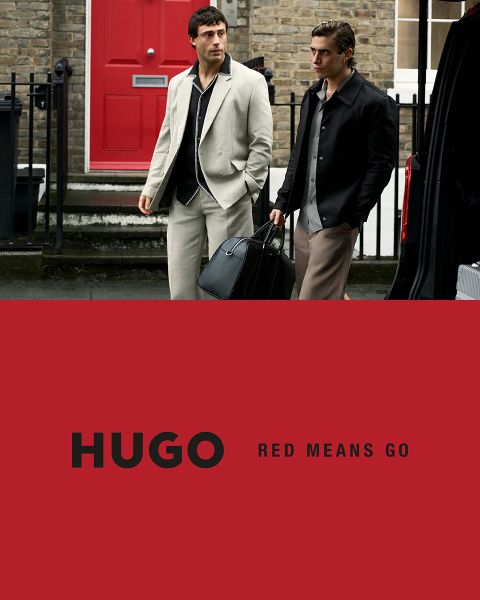 Dos modelos en trajes negros posan con confianza frente a un fondo claro y sencillo. El logo de HUGO aparece en letras blancas y gruesas en la esquina inferior izquierda.