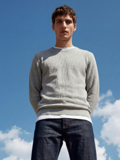 Un joven que lleva una camiseta negra con la inscripción "Original Studio", un collar plateado, una pulsera plateada y una gorra de béisbol a rayas color crema y negro, está de pie frente a un fondo liso de color gris claro.