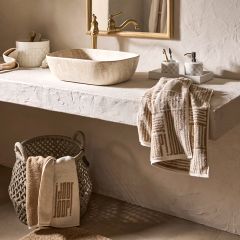Baño minimalista con espejo redondo, grifo de pared y lavabo rectangular. Estantes sostienen elementos decorativos y toallas en tonos tierra. La luz suave proyecta sombras delicadas sobre la pared texturizada, creando un ambiente tranquilo.