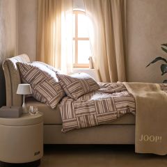 Un dormitorio acogedor con una cama beige con ropa de cama estampada. Un espejo redondo cuelga en la pared. Una luz suave ilumina una parte de la cama. Una pequeña pila de libros y objetos de decoración se encuentran cerca en el suelo.
