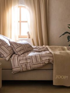 Un espacio minimalista presenta un banco de piedra beige con dos cojines marrones y beige texturizados, una manta estampada y hierbas secas. Una cesta trenzada, un jarrón de cerámica rústica y una jarra alta adornan el suelo bajo una suave luz natural.