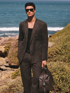 Una persona con un traje negro formal está de pie frente a una barandilla de piedra con árboles al fondo. El cielo está parcialmente nublado. Su camisa está parcialmente desabrochada y tiene una expresión facial segura de sí misma.