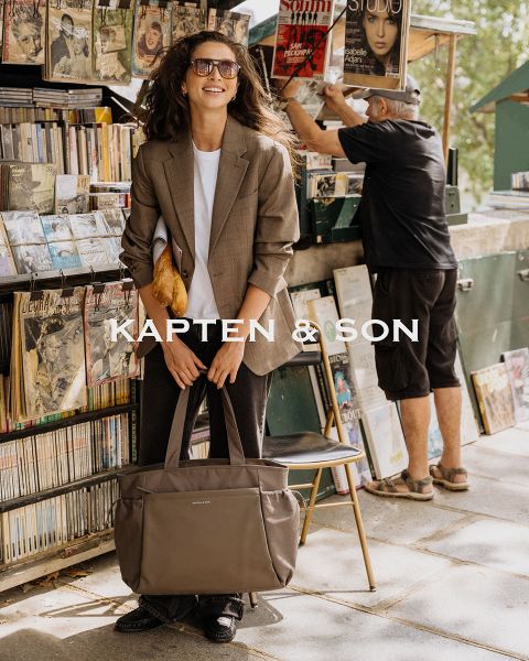 Una mujer y un hombre se sonríen al aire libre. La mujer lleva un gorro verde y un abrigo beige y sostiene un bolso negro en la mano. El hombre lleva una chaqueta marrón y un bolso color crema. En el centro se lee "Kapten & Son".