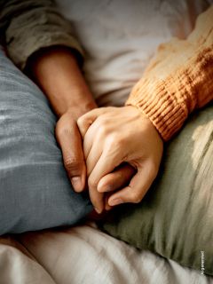 Dos personas acostadas en una cama tomadas de la mano, una con una manga naranja y la otra con una manga verde, transmitiendo consuelo y cercanía.