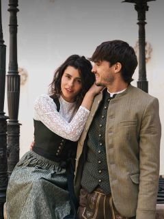Una mujer y un hombre con trajes bávaros tradicionales están sentados juntos al aire libre, sonríen y se miran.