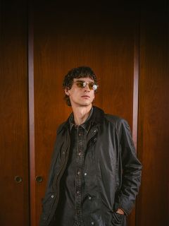 Una persona con gafas de sol y chaqueta oscura está de pie frente a puertas de madera, mirando ligeramente hacia un lado y con las manos en los bolsillos.