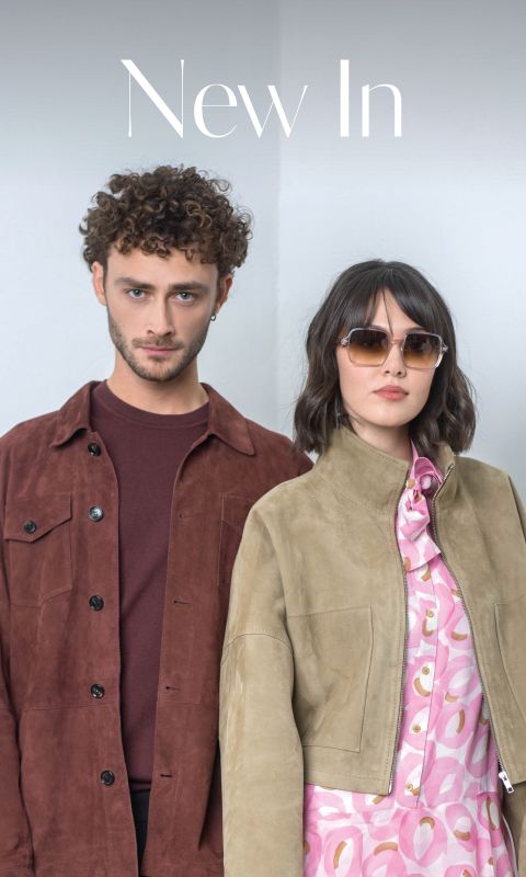 Un hombre con una chaqueta marrón y una mujer con gafas de sol y una chaqueta marrón claro sobre un vestido con estampado rosa están de pie uno al lado del otro frente a un fondo claro y sencillo.