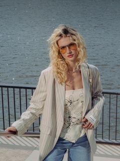 Una mujer con cabello rubio rizado lleva gafas de sol, una chaqueta a rayas, una blusa floral, pantalones cortos de mezclilla y zapatos blancos mientras está de pie en un paseo marítimo con barandilla.