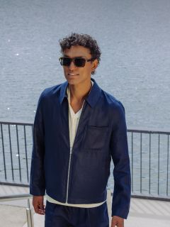 Un hombre con gafas de sol, chaqueta azul marino y pantalones cortos a juego sube una escalera exterior junto a un cuerpo de agua, agarrándose a una barandilla y sonriendo.