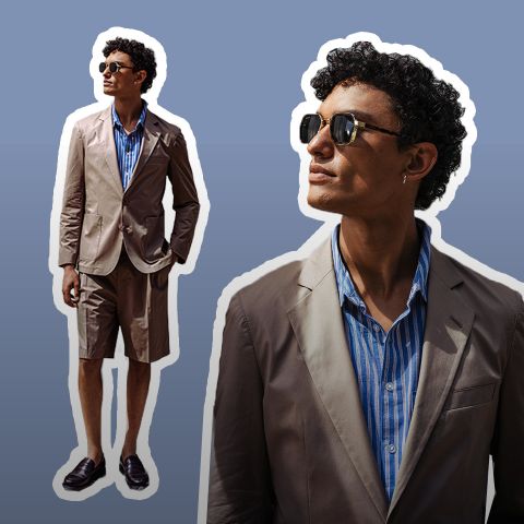 Hombre con gafas de sol, blazer tostado, camisa de rayas azules y pantalones cortos tostados, de pie con confianza sobre un fondo degradado marrón.