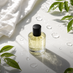 Un frasco de perfume transparente con tapa negra está sobre una superficie blanca con gotas de agua, hojas verdes y una toalla blanca cerca.