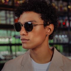 Una persona con pelo corto y rizado, que lleva gafas de sol, una chaqueta beige y una camisa blanca, está de pie en un interior con estantes de vidrio al fondo.