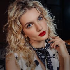 Una mujer con cabello rubio rizado, lápiz labial rojo y una bufanda de lunares mira a la cámara y se lleva la mano a la nuca.