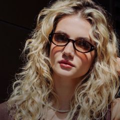Mujer joven con cabello largo, rizado y rubio, que lleva gafas de carey, un collar de perlas y una blusa marrón, mirando a la cámara.