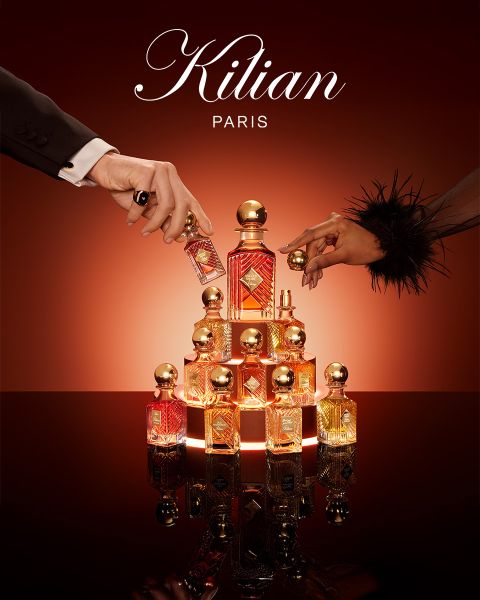 Un frasco del perfume Kilian Paris Angels' Share está junto a un vaso de whisky con hielo y una guarnición, ambos sobre una superficie marrón reflectante con iluminación suave. Arriba se ve el logo de Kilian Paris.