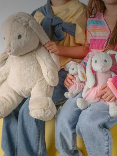 Un niño sonriente con cabello rubio está sentado en una casa abrazando a un gran oso de peluche beige. Lleva una camisa de manga larga blanca y negra y pantalones claros.