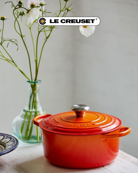 Una escena de cocina con una olla de cocción lenta roja, una sartén antiadherente, un juego de ollas apilables de acero inoxidable, una olla negra y un cuenco de tomates cherry sobre una encimera de madera clara frente a un protector contra salpicaduras de azulejos blancos. El logotipo de Le Creuset es visible.