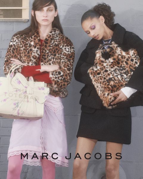 Una mujer posa al aire libre con un bolso marrón de Marc Jacobs en la mano, se inclina hacia atrás con la cabeza ladeada y lleva una chaqueta color castaño y vaqueros azules. En la imagen aparecen las palabras "EL BOLSO TOTE MARC JACOBS" y "MARC JACOBS".