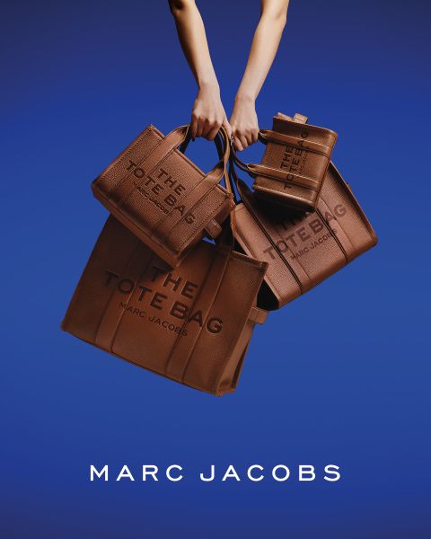 Una mujer posa al aire libre con un bolso marrón de Marc Jacobs en la mano, se inclina hacia atrás con la cabeza ladeada y lleva una chaqueta color castaño y vaqueros azules. En la imagen aparecen las palabras "EL BOLSO TOTE MARC JACOBS" y "MARC JACOBS".