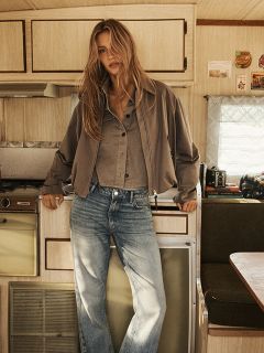 Una mujer con cabello rubio largo y ondulado lleva una capa marrón con capucha y un cinturón negro, está de pie frente a una pared rústica de madera y mira con confianza a la cámara.